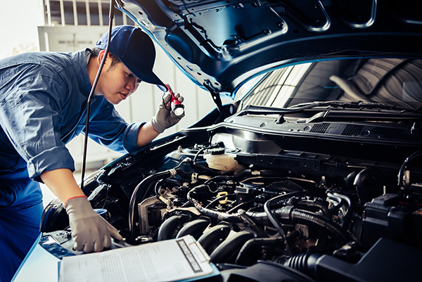 Car mechanic technician holding flashlight checking engine with checklist clipboard for pre-purchase inspection.