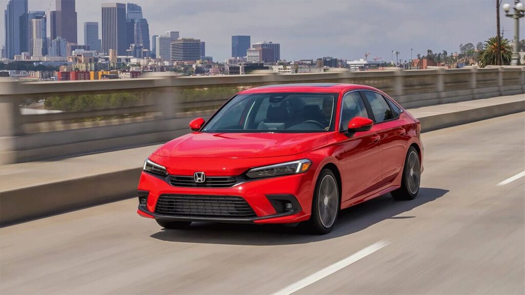 A red Honda Civic sedan driving on a freeway with buildings visible in the distance.